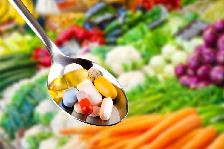 vitamíny v tabletkách na lyžičke, v pozadí množstvo čerstvej zeleniny