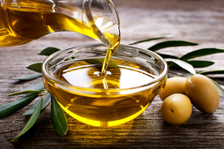 olivový olej niekto leje z čaše do misky, vedľa je sú olivy a listy z olivovníka