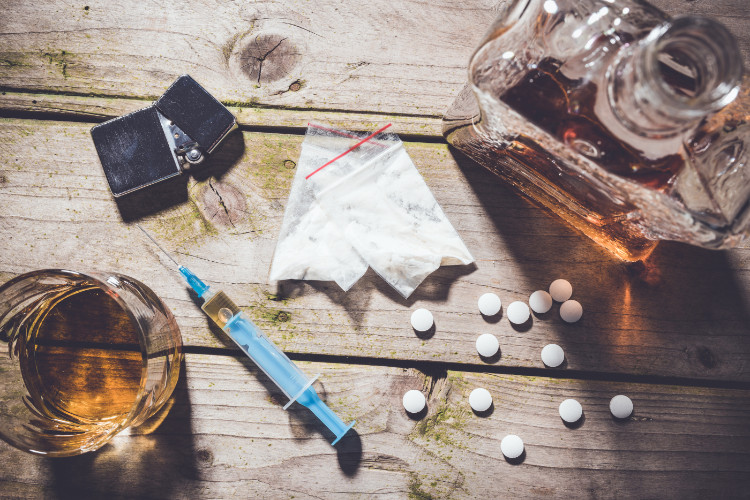 na drevennom stole je sklenený pohár a fľaša s alkoholom, rozsypané lieky, injekčná striekačka, vrecúška s drogami a zapaľovač