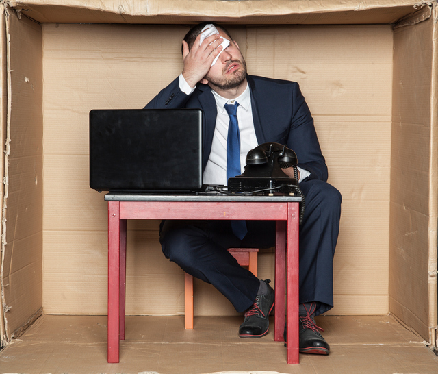 Biznismen sedí za stolom v krabici, stiesnené priestory, pracovný stres a zvýšená záťaž, počítač a telefón, muž si utiera čelo