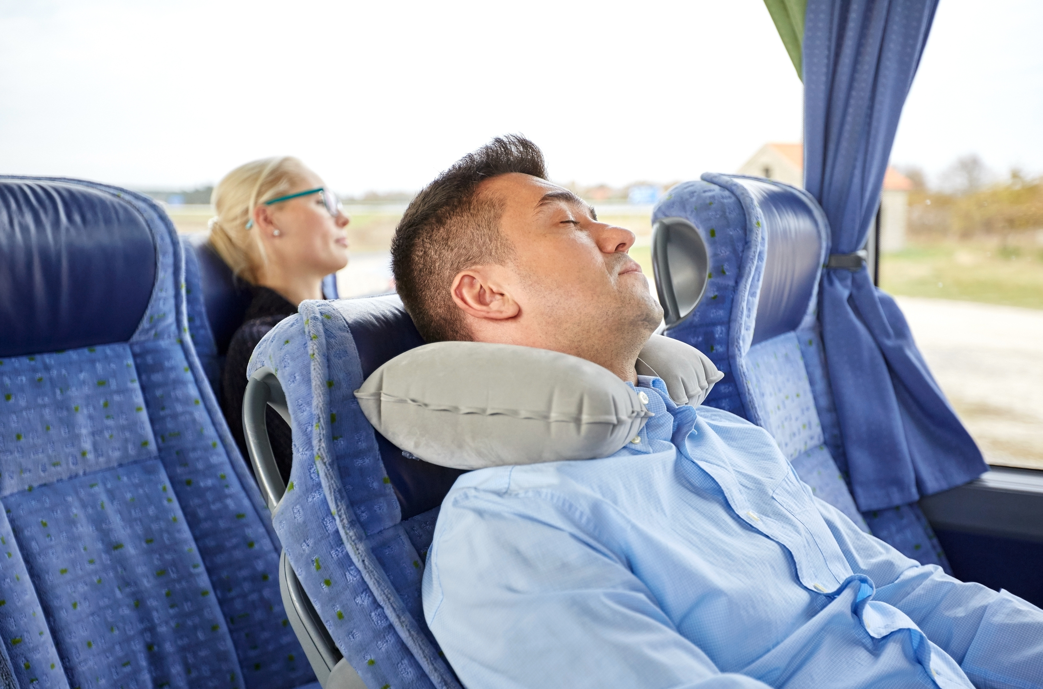 Dvaja cestujúci, spiaci v autobuse, muž má vankúš. Sedadlá, vonku cez okná vidieť stromy