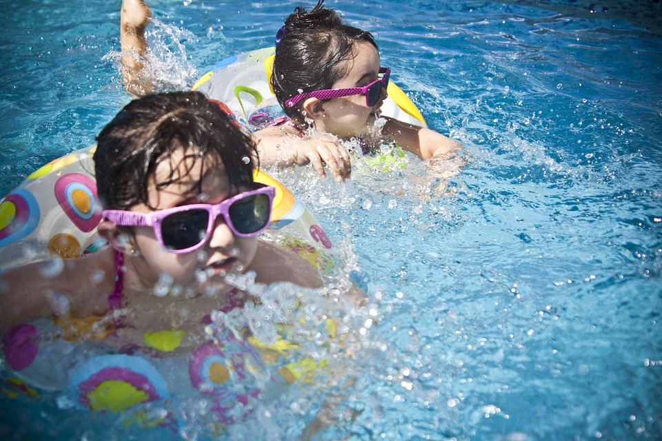 Dve deti sa kúpu vo vode. Majú slnečné okuliare s ružovým rámom. Sú v plávacích kolesá.
