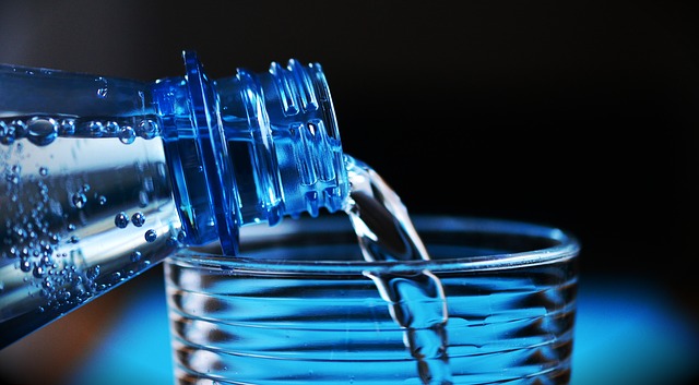 Voda sa nalieva s fľaše do pohára. Vidno iba hrdlo fľaše a vrch pohára. Pozadie je tmavé a modré.