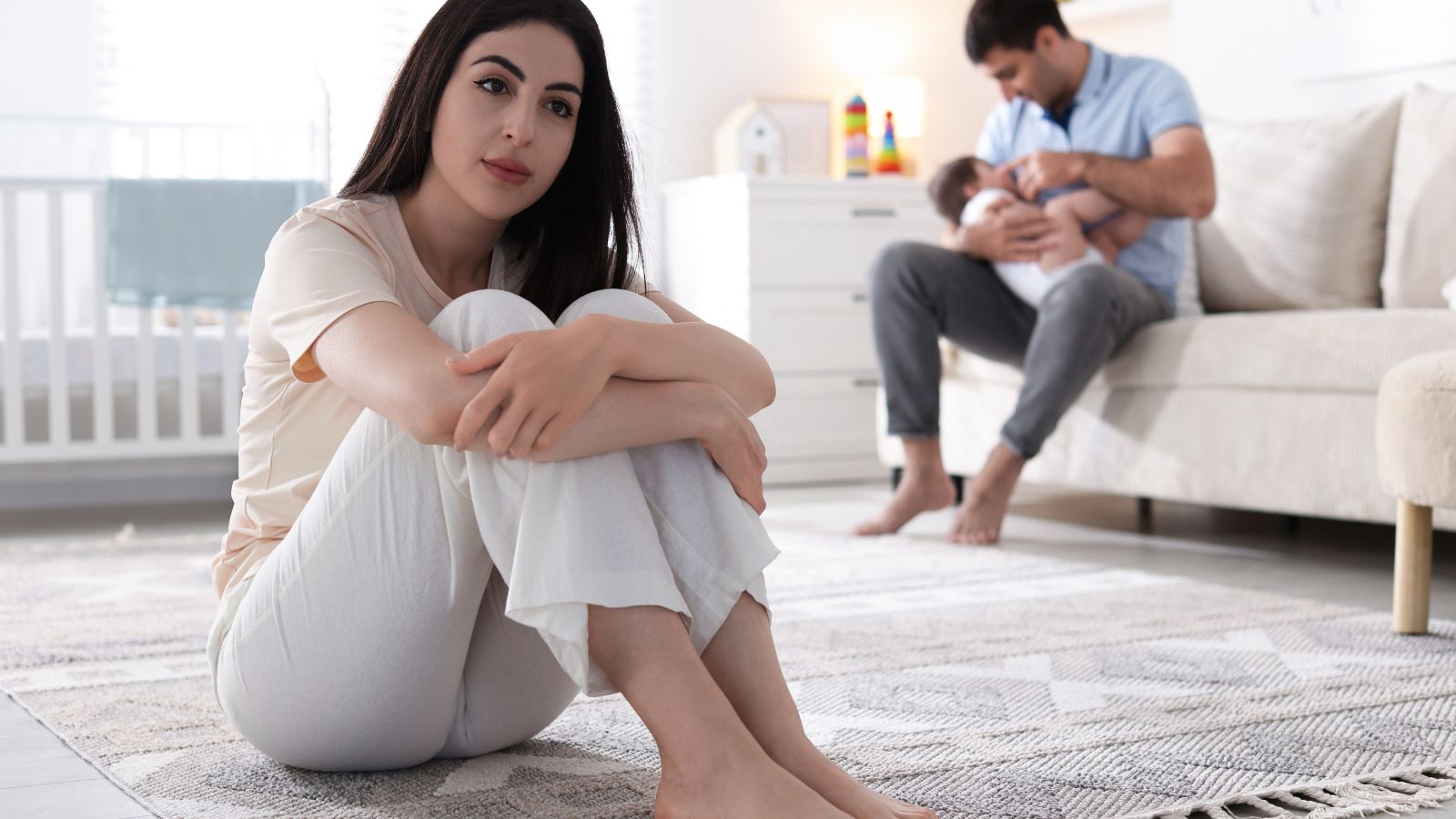 Matka sedí sama na koberci v detskej izbe. V pozadí na pohovke sedí jej partner s dieťaťom v náručí.