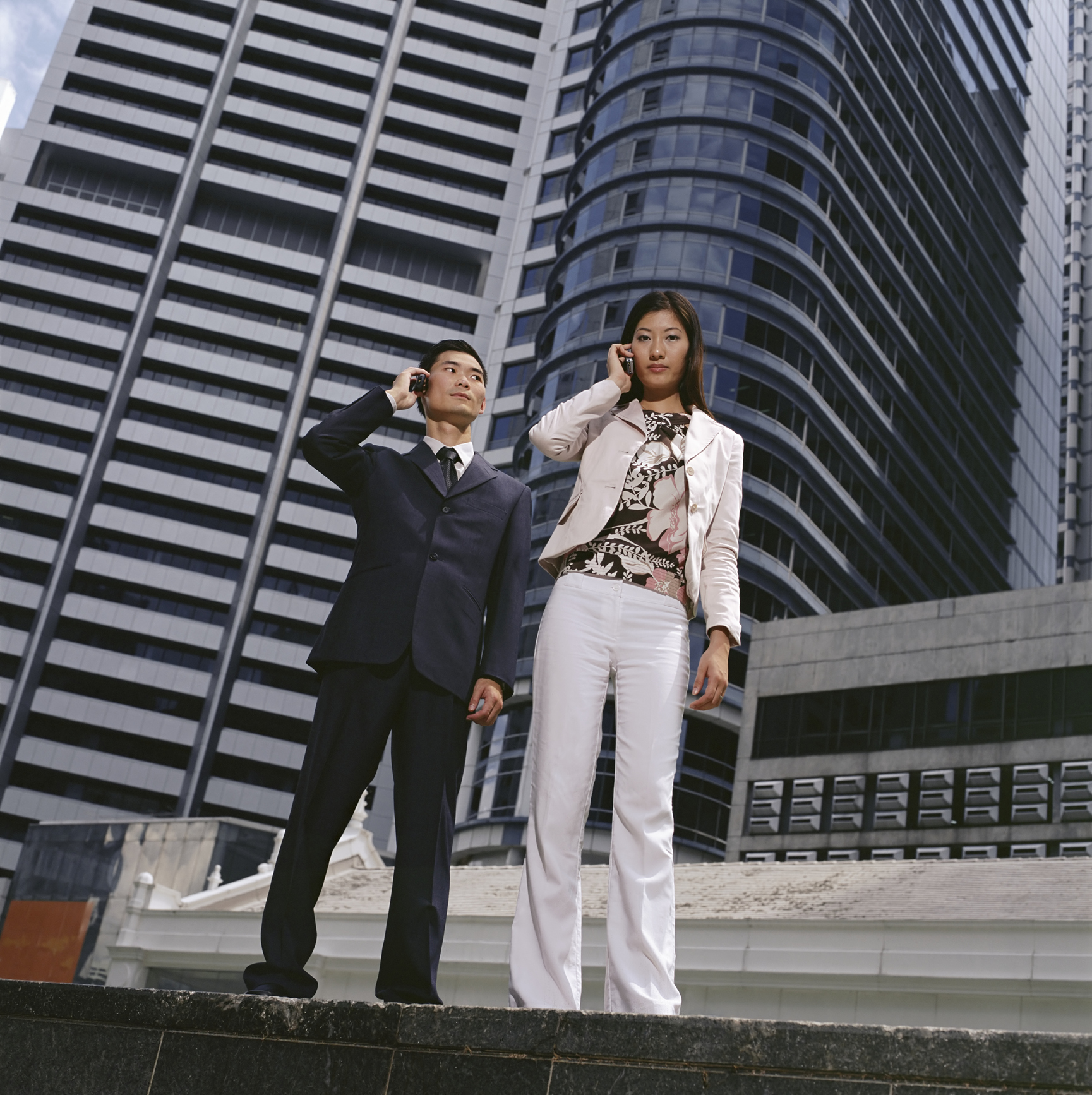 Žena a muž telefonujú z mobilov, v pozadí výškové budovy.