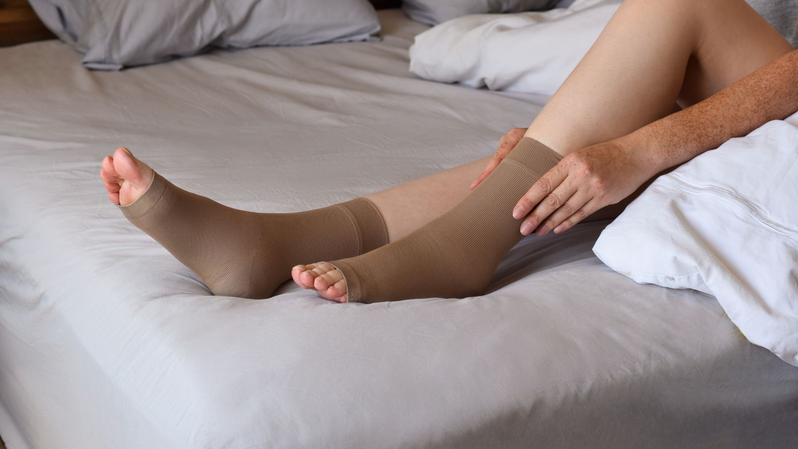 V lete má naše telo väčšiu tendenciu zadržiavať vodu - vysoké teploty, nadmerný príjem soli, sedavý spôsob života, hormonálne zmeny, či dokonca niektoré lieky môžu prispieť k zadržiavaniu vody v tele. Zdroj foto: Canva Na posteli sú vystreté nohy zobrazené od predkolenia. Na nohách sú oblečené kompresné ponožky s voľnou špičkou.