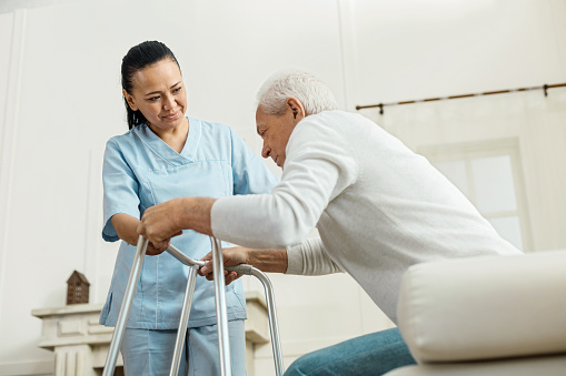 starší mž sediaci na nemocničnej posteli sa snaží postaviť za pomoci chodítka a zdravotnej sestry