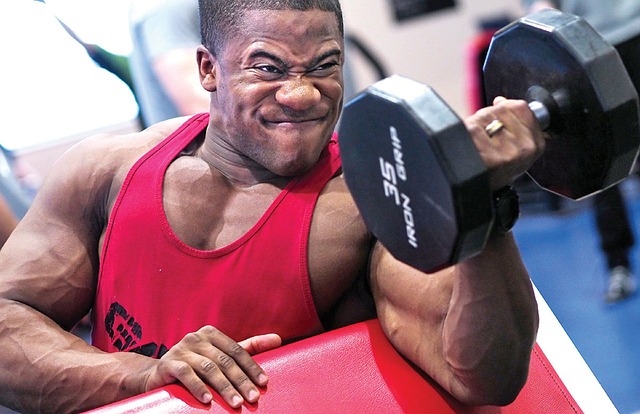 Muž v červenom tielku. Posilňuje biceps s jednoručnou činkou.
