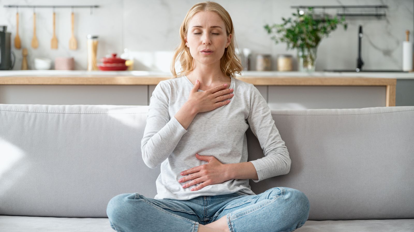 Dychové cvičenia sú techniky zamerané na vedomé riadenie a kontrolu dýchania s cieľom ovplyvniť telesné a psychické funkcie. Zdroj foto: Canva Žena sediaca na pohovke v tureckom sede. Pravá ruka položená na hrudníku, ľavá na bruchu. Kontrola vedomého dýchania, relaxácia, uvoľnenie úzkosti.