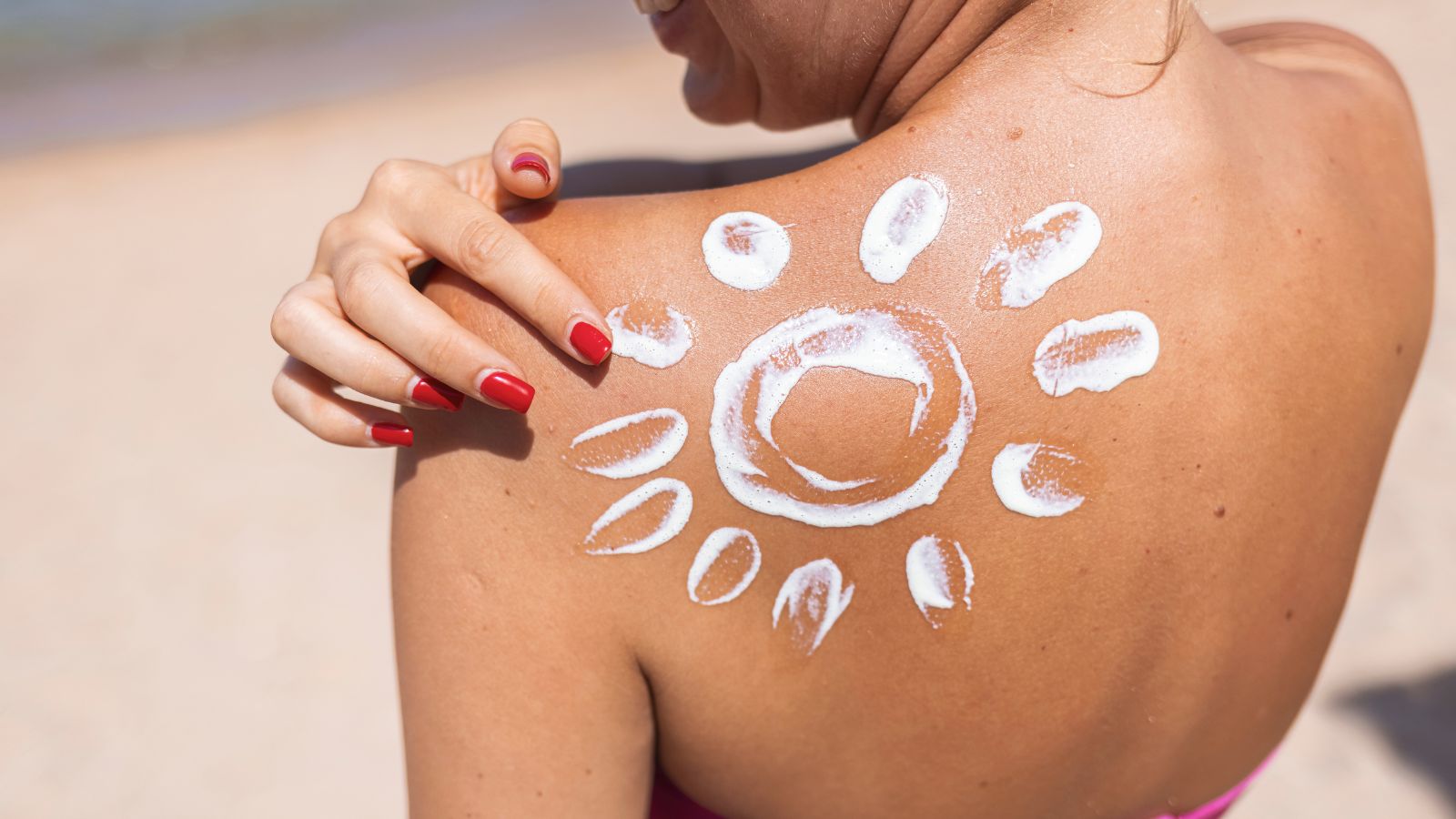 Žena s opálenou pokožkou. Záber hornej časti chrbta. Aplikuje krém s SPF ochranou, ktorý je natrtý do tvaru slnka na lopatke. Bezpečné opaľovanie.