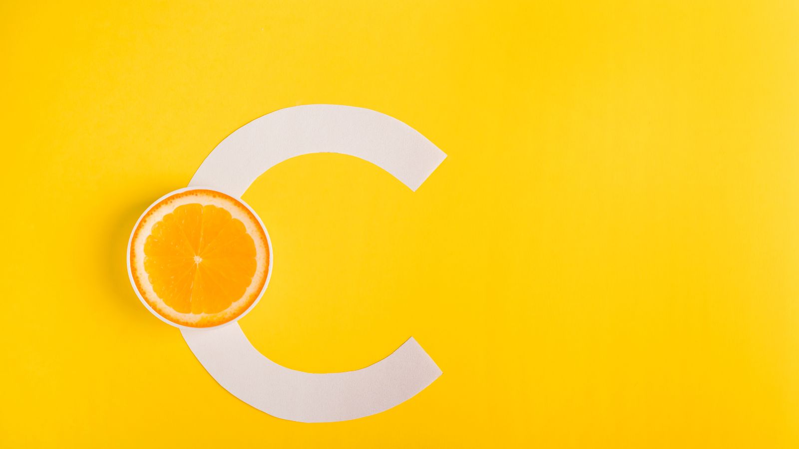 obrázok so žltým pozadím s bielym písmenom C, cez ktoré je položený plátok pomaranča. Symbol vitamínu C.