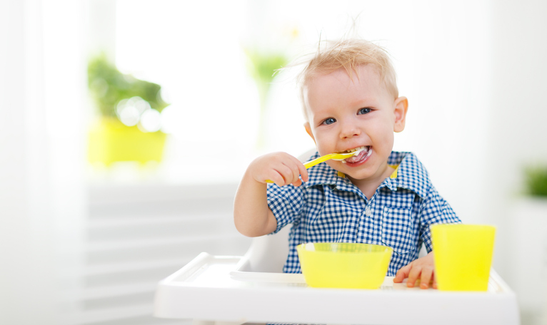 Malé dieťa sedí v detskej sedačke, kŕmi sa. Žltá lyžička, pohár a miska